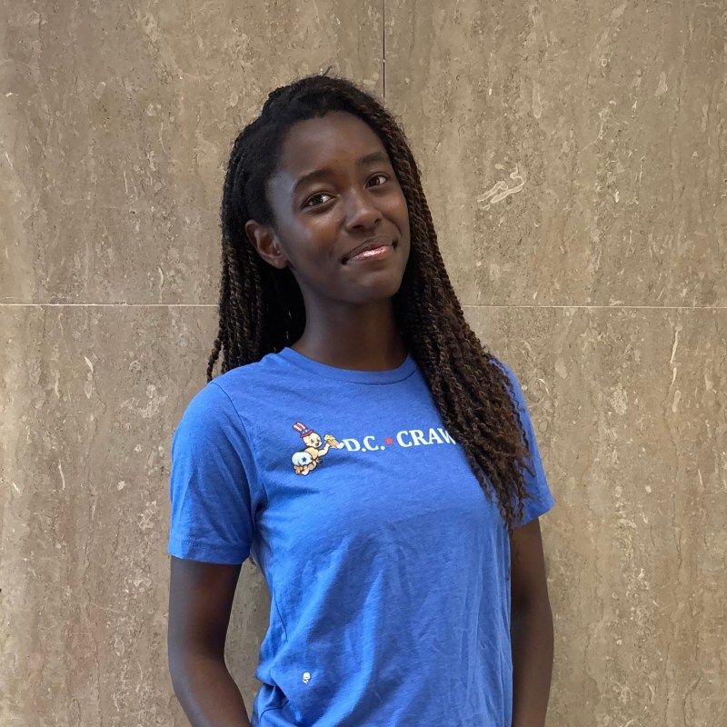 Smiling person in blue T-shirt with 'D.C. Crank' text, standing against a beige marble wall.